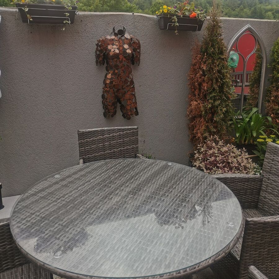 A round glass-topped wicker table in the courtyard in front of a grey wall with flower boxes and a copper-coloured wall sculpture