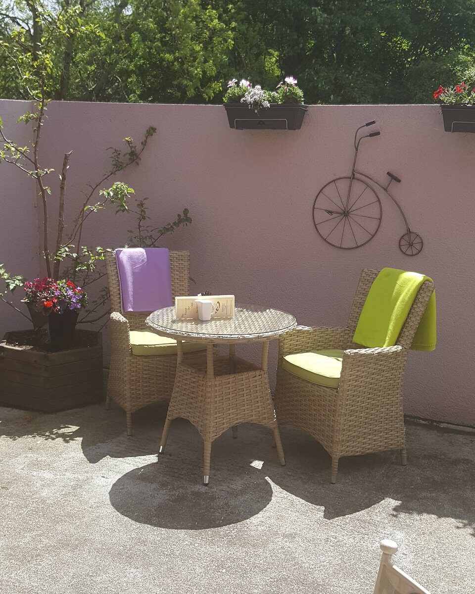 Two wicker chairs with green cushions and a small table in the sunny courtyard, in front of a pink wall with a bicycle ornament